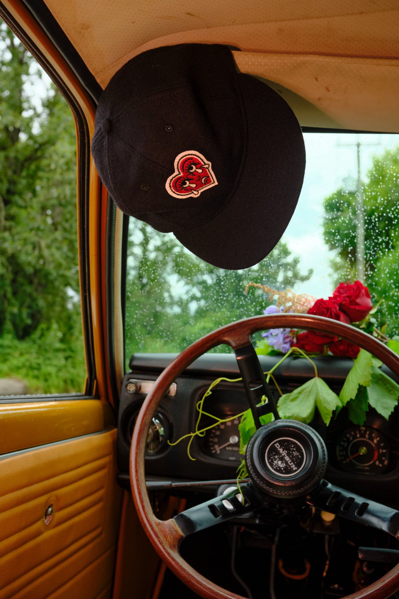 Black cap with a crying heart logo patch on a car dashboard with flowers and steering wheel visible