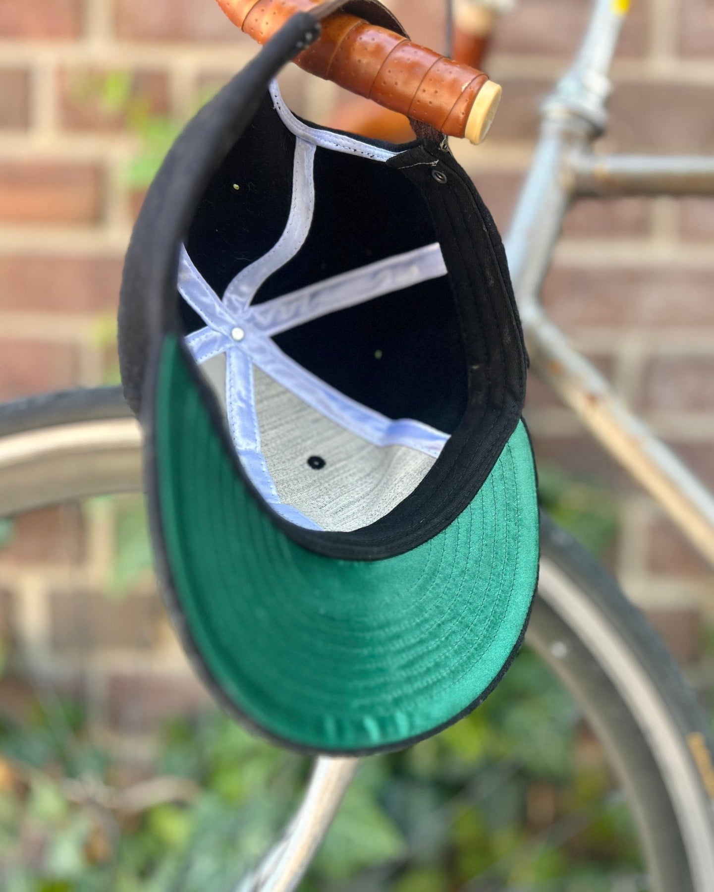 Black cap with a green under-brim hanging on a bicycle against a brick wall.