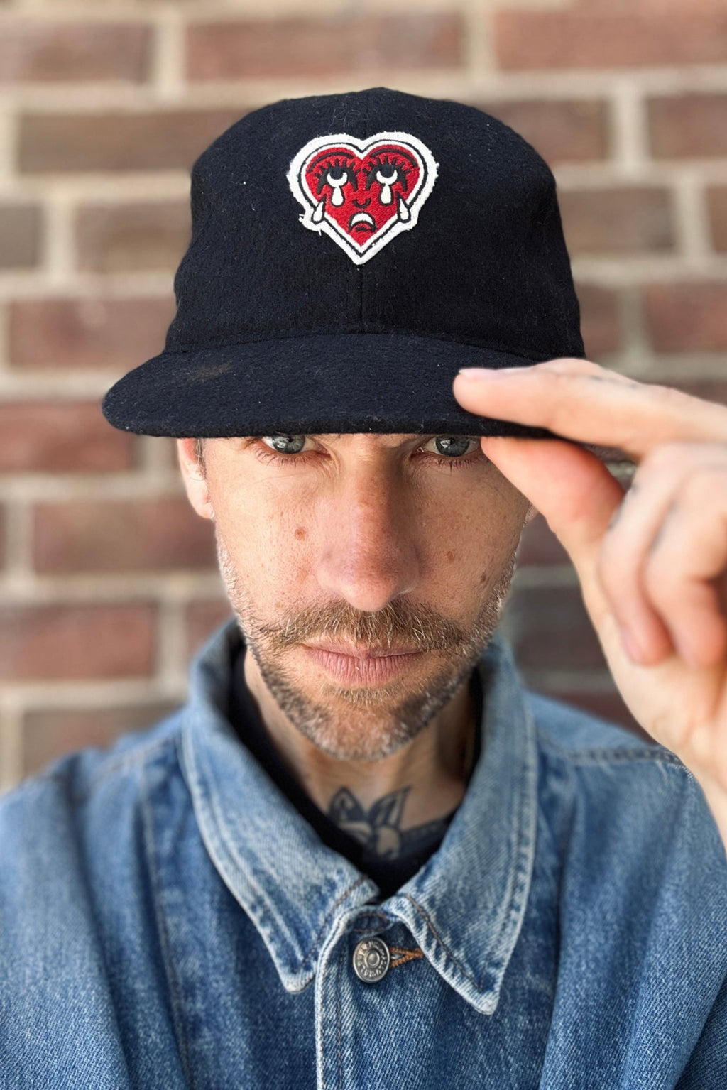 Man wearing a black cap with a crying heart patch against a brick wall