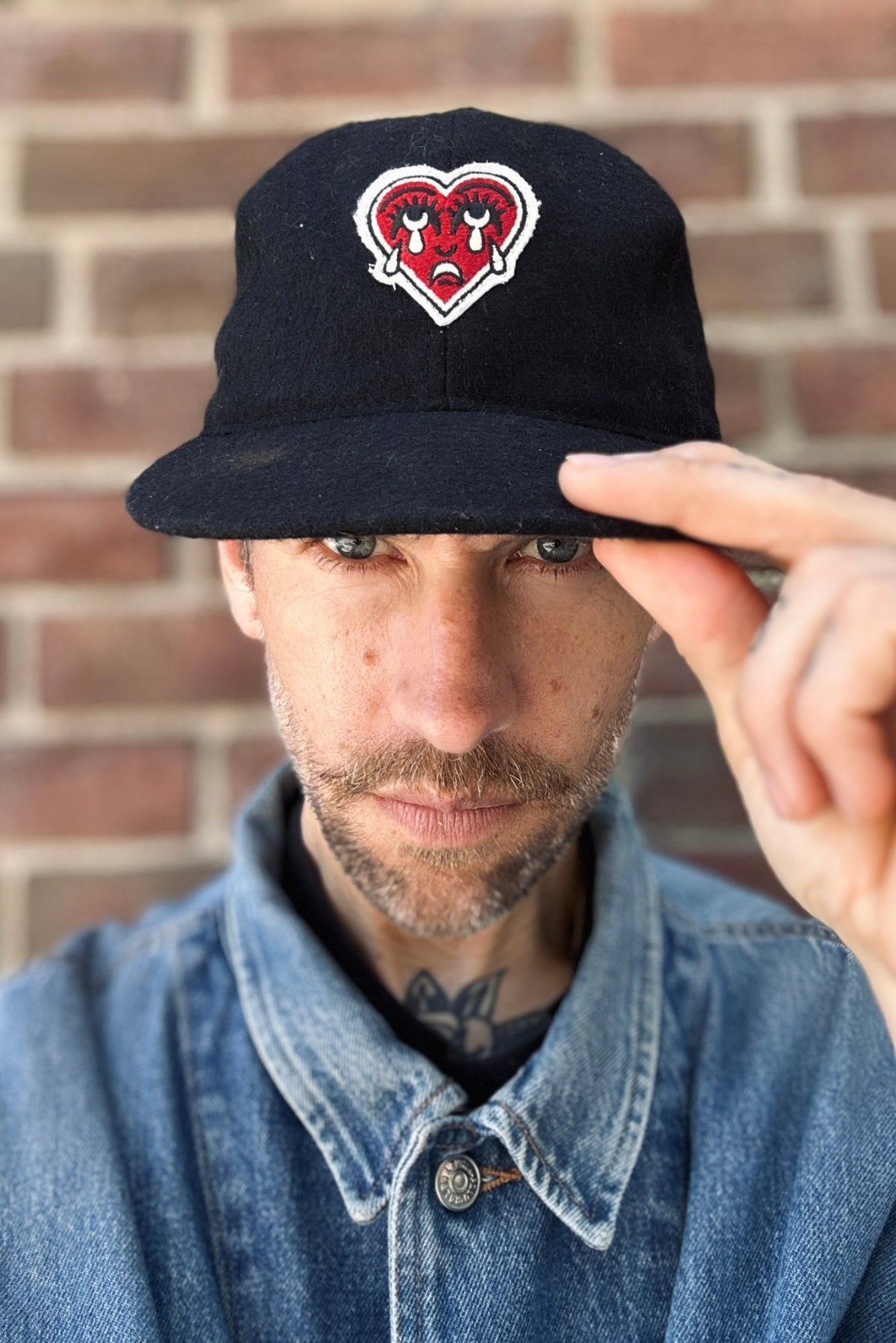 Man wearing a black cap with a crying heart patch against a brick wall