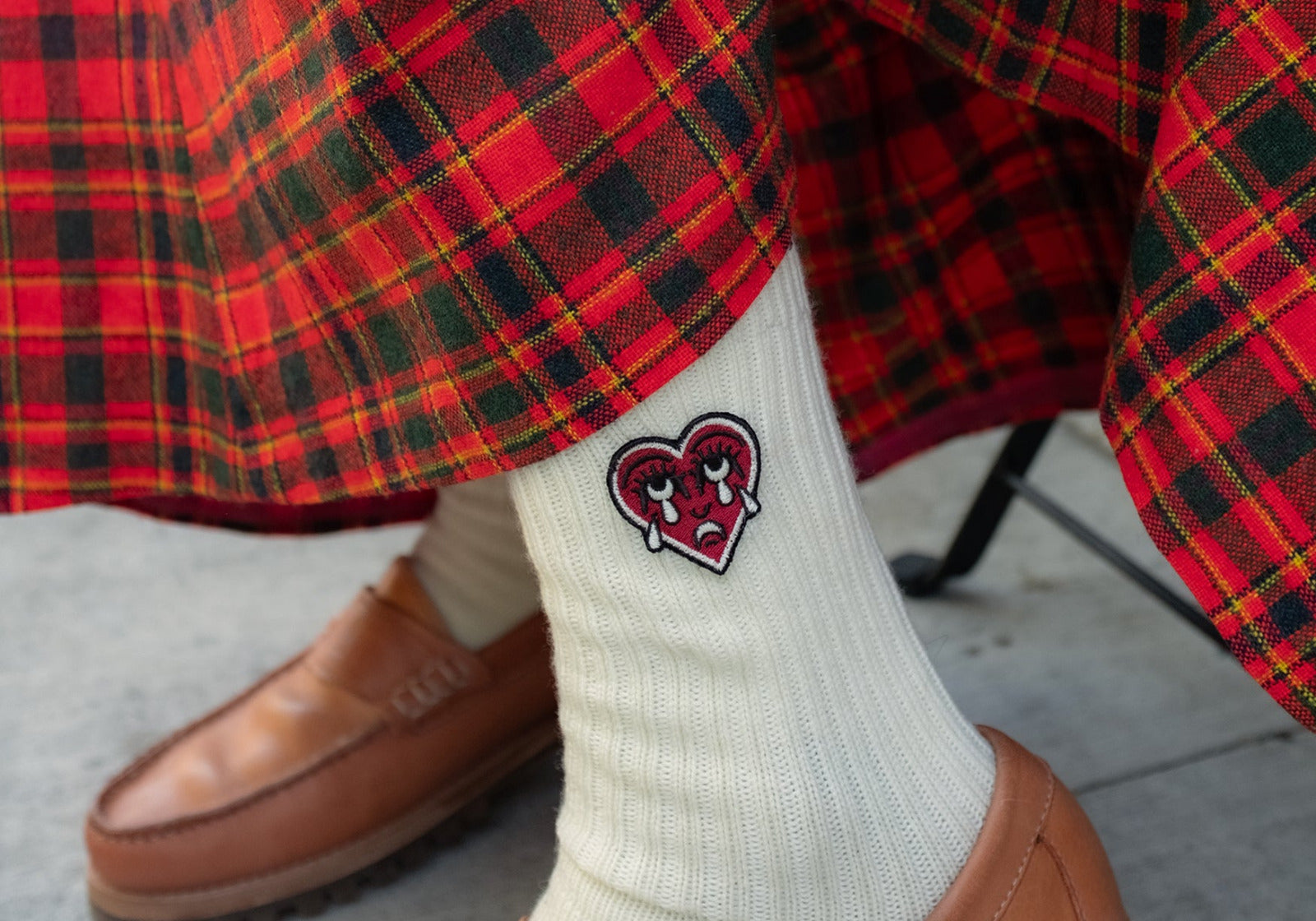 White sock with a red crying heart logo worn with a plaid skirt and tan shoes.