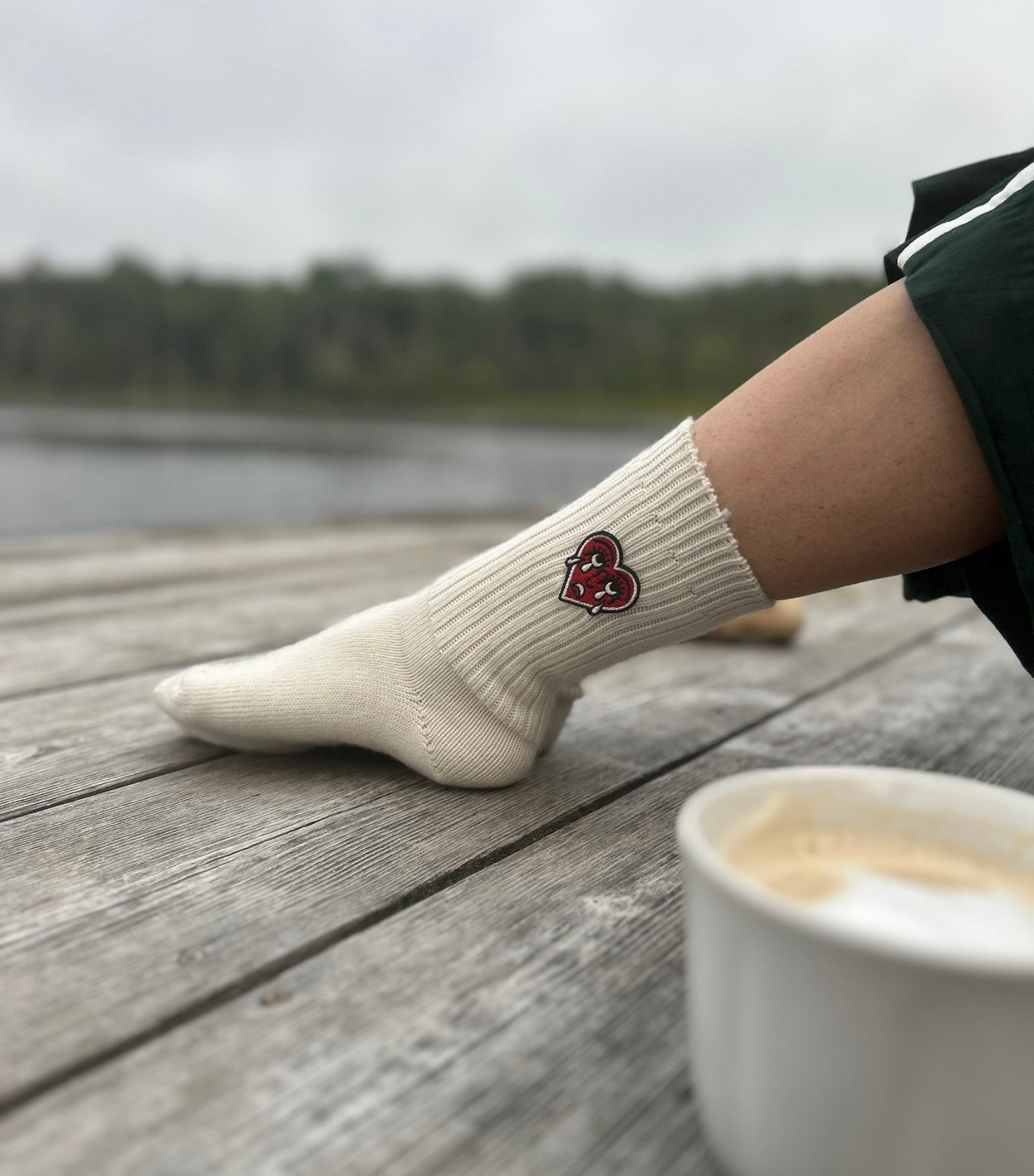 Person wearing a natural sock with a red crying heart logo on a wooden dock by a lake.